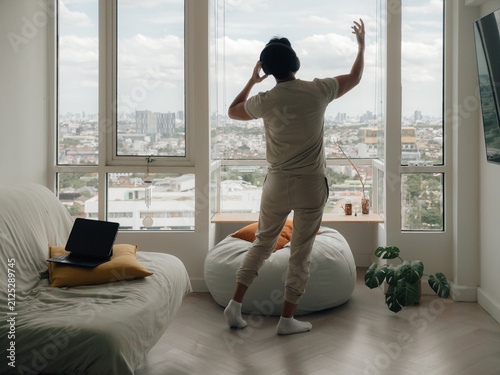 Relaxed man in headphones dancing by a window, enjoying music and city views at home.