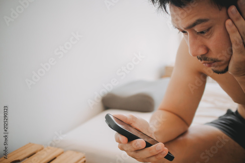 Serious Asian Man Checking His Mobile Phone in Bed, Looking at Screen with Concern