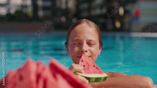 A smiling boy stands in a swimming pool holding a slice of fresh watermelon on a hot summer day. Wet from swimming, he enjoys a healthy fruit snack while relaxing at an outdoor resort pool. The bright