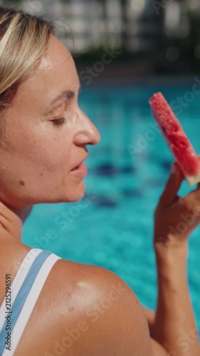 Young woman sitting by an outdoor swimming pool on a sunny summer day, enjoying a fresh slice of watermelon. She takes a bite of the juicy fruit while relaxing poolside, creating a carefree vacation