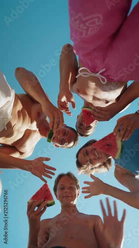 Low-angle view of a family standing in a circle outdoors on a sunny summer day, holding fresh watermelon slices and enjoying a healthy snack together. The camera looks up toward the blue sky, creating