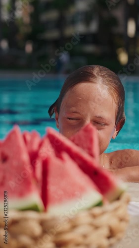 A smiling boy stands in a swimming pool holding a slice of fresh watermelon on a hot summer day. Wet from swimming, he enjoys a healthy fruit snack while relaxing at an outdoor resort pool. The bright