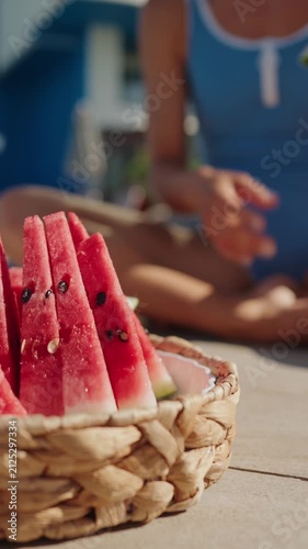 Close-up of a woven basket filled with fresh watermelon slices by an outdoor swimming pool on a sunny summer day. A woman in a blue swimsuit reaches for a slice, creating a relaxed vacation atmosphere