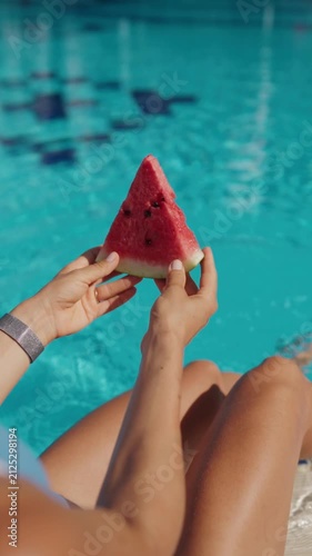 A woman relaxes by an outdoor swimming pool holding a fresh slice of watermelon on a sunny summer day. Viewed from behind, she enjoys a peaceful vacation moment while cooling her feet in the water