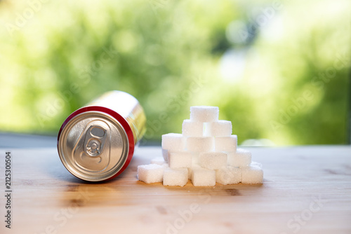 A can of a sweet soft drink next to a stack of sugar cubes on a kitchen counter. Blurred trees in the background. Sweet food, white sugar, sweet drink, non-alcoholic beverage.