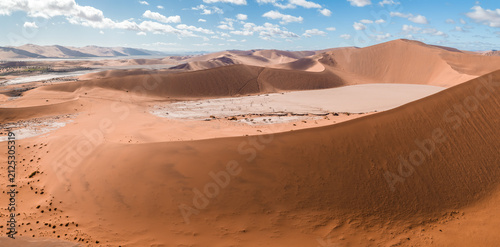 Canvas Print Aerial view shows red dunes of Sossusvlei around white Deadvlei pan in Namibia, with tiny hikers on ridge lines under cumulus clouds and warm light