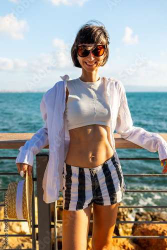 Sensual girl with piercing and sunglasses poses in the sun with dark hair fluttering in the wind. Calm brunette stands against golden evening sunset.
