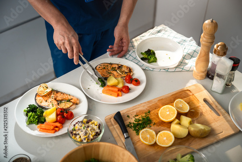 Healthy Eating with a Caregiver Arranging Grilled Salmon, Broccoli, Carrots, Potatoes and Roasted Tomatoes on Plates in a Bright Home Kitchen