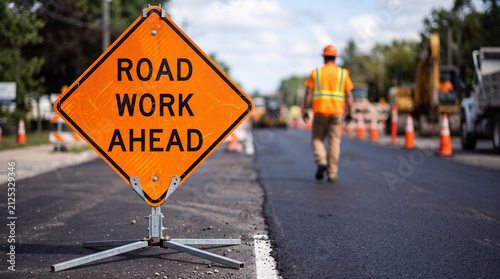 A close-up of a road work ahead sign on a newly paved road, with a construction man walking in the background. Concept of road construction, infrastructure repair.