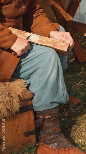 A person in medieval clothing carves a piece of wood while seated on the ground. The individual is focused on their work, surrounded by simple wooden tools and natural materials