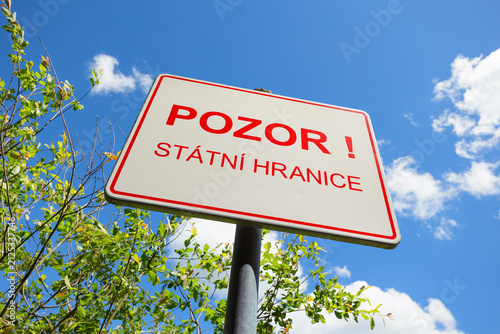 Czech green border warning sign with text Pozor! Státní hranice (translation: Attention! State Border). Sunny with clear blue sky and green vegetation. Copy space, shallow focus, low angle view.