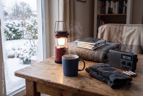 A setup includes a red lantern, thermal blankets, gloves, and a plain mug with a hot drink. Snow covers the ground outside a window. A radio sits beside the items