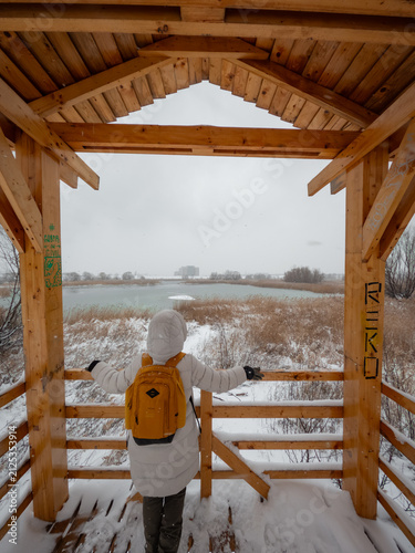 Fotografía Rear view of a female hiker in a white coat and yellow backpack standing in a wooden observation tower, admiring a snow-covered landscape and frozen lake in Vacaresti Nature Park, Bucharest, Romania