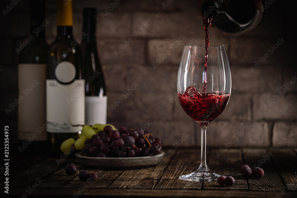 Obraz premium A dark, elegant still life of red wine being poured into a glass with wine bottles and fresh grapes in the background. Perfect for wine-themed compositions