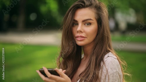 Beautiful attractive young woman texting on smartphone in green park, then looking at camera. Pretty female holding mobile phone on blurred nature background outdoors.