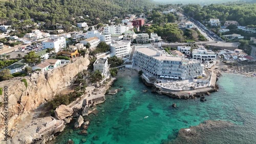 Aerial forward push-in shot flying over turquoise ocean toward resort hotels and coastal buildings in Cala Sant Vicenc, Mallorca