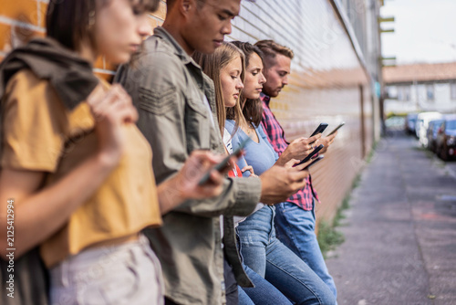 Obraz na plátně Multiethnic gen z friends stand together while absorbed in smartphones beside an urban wall during an outdoor meetup