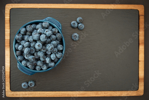 Top view of fresh, ripe blueberries filled in a blue ceramic bowl placed on a black slate serving board with wooden frame, leaving copy space on the right.