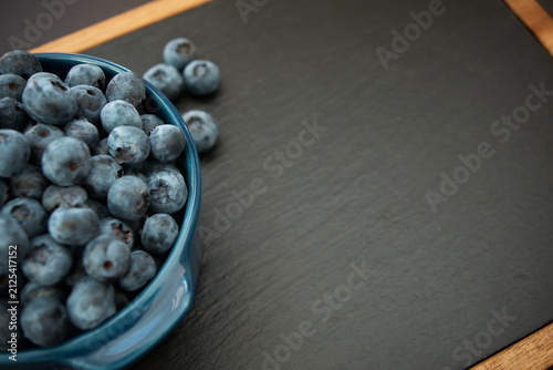 Top view of fresh, ripe blueberries filled in a blue ceramic bowl placed on a black slate serving board with wooden frame, leaving copy space on the right.