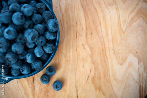 A top view close-up of a blue ceramic bowl overflowing with fresh, ripe blueberries on a natural light wood background with copy space.