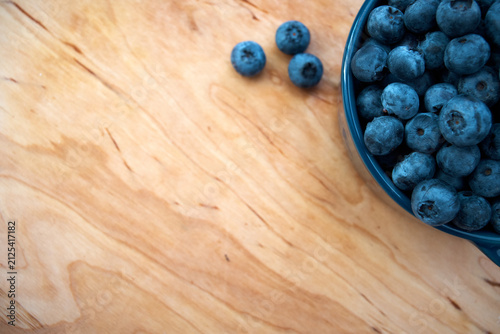 A top view close-up of a blue ceramic bowl overflowing with fresh, ripe blueberries on a natural light wood background with copy space.