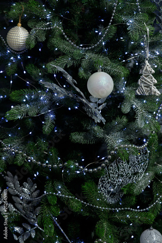 Green Christmas tree with white and silver baubles and glittery leaves Silver bead garlands wrap branches with LED lights glowing in dark background creating festive holiday mood