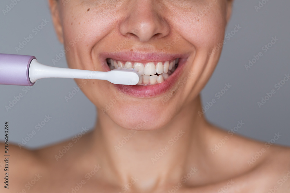 Fototapeta premium Close-up of young woman brushing teeth with electric toothbrush, smiling during morning oral hygiene routine on gray background
