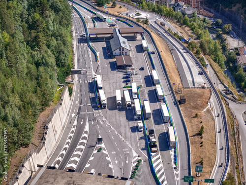 Aerial view of the Traforo del Frejus entrance with trucks waiting in lanes at the toll plaza surrounded by dense forest and mountain terrain in Bardonecchia, Piemonte, Italy.