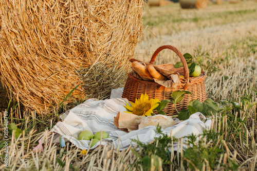 A woven basket filled with bread and apples sits beside a golden hay bale, with a sunflower and rustic blanket completing the countryside scene.