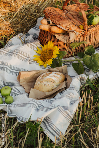 A woven basket filled with bread and apples sits beside a golden hay bale, with a sunflower and rustic blanket completing the countryside scene.
