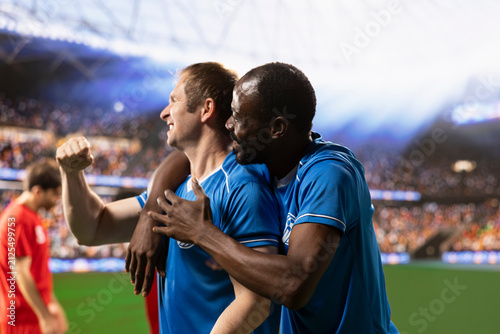 фотографія Diverse football players celebrate a winning goal with raised arms and clenched fists as teammates hug and defeated opponents remain on the pitch