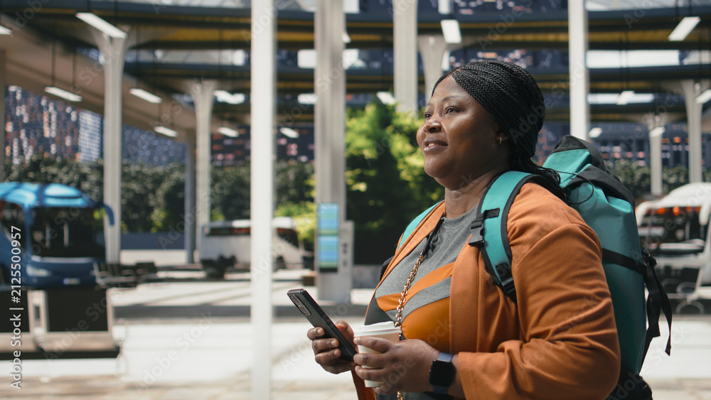 Fototapeta premium Woman enjoys coffee and uses a smartphone in a bus station, carrying a takeaway drink and checking travel information for an intercity coach departure in a close-up scene as a solo passenger.