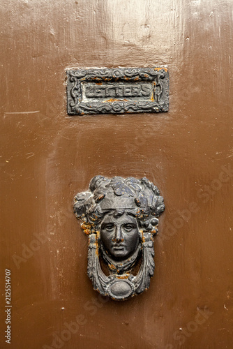 An ornate bronze door knocker shaped like a face sits beneath a vintage letterbox on a brown door, Malta