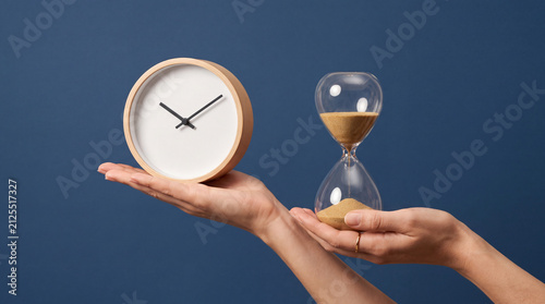 A woman holding a wooden wall clock and a glass hourglass, symbolizing time management, productivity, and the passing of time against a blue background.