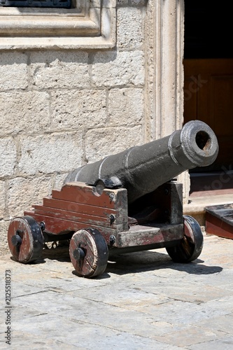 Konstfotografi Close-up of an ancient black cast-iron cannon mounted on a brown wooden carriage with wheels, standing outdoors against a vintage stone fortress wall