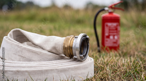 A rolled-up white industrial fire hose with a metal coupling placed on the grass next to a red fire extinguisher, prepared for safety and emergency response.