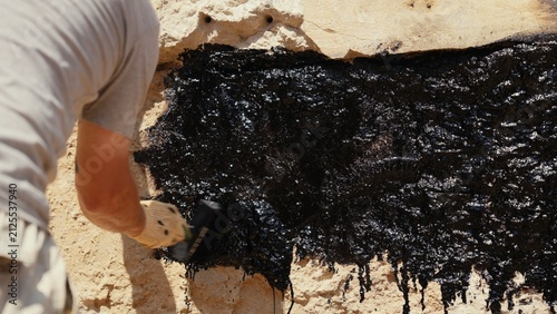 Construction worker applying black liquid sealant to a stone wall surface, creating a protective barrier against moisture and water damage for building foundations