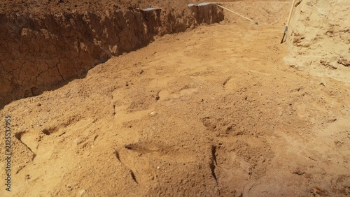 Excavated earth forming a construction trench, revealing sandy soil layers with cracked clay walls and visible human footprints on the ground, indicating recent activity