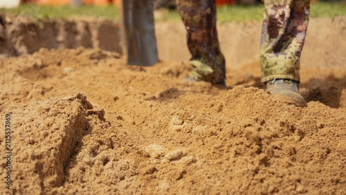 Fotografía Person wearing camouflage pants and work boots standing in a trench, using a sho