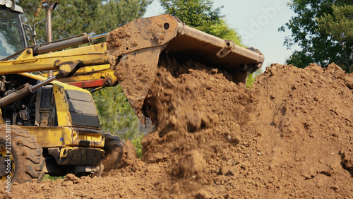 Yellow backhoe loader with a large bucket filling a pile of soil, performing excavation work on a construction site under a clear sky