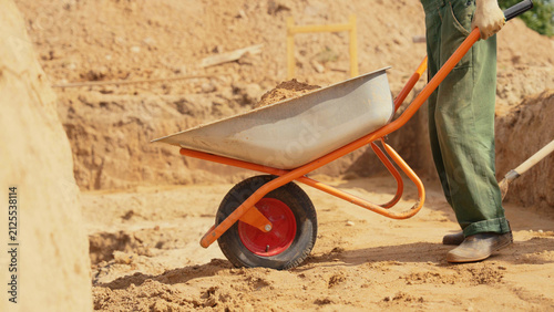 Worker pushing an orange wheelbarrow filled with soil, performing manual labor for excavation and building groundwork on a sunny construction site