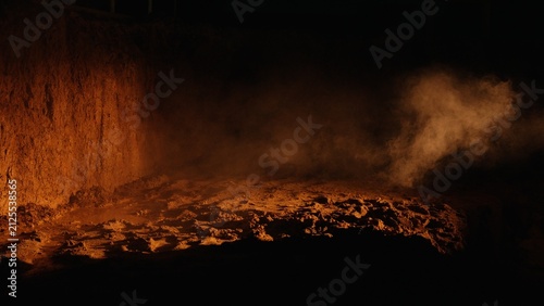 Underground excavation showing a dark pit with textured clay and soil illuminated by a mysterious orange light, creating a dramatic atmosphere with swirling smoke and fog