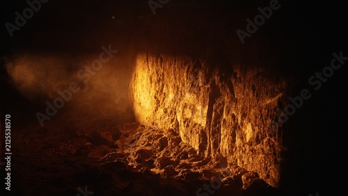 Dark cavern showing a rough earth wall illuminated by a golden light source, with smoke wafting through the deep shadows, creating an atmospheric and textured subterranean scene
