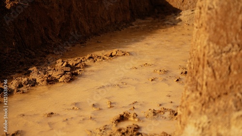 Earthwork showing a narrow trench filled with brown, silty water and wet dirt, creating a challenging and messy path after heavy rain