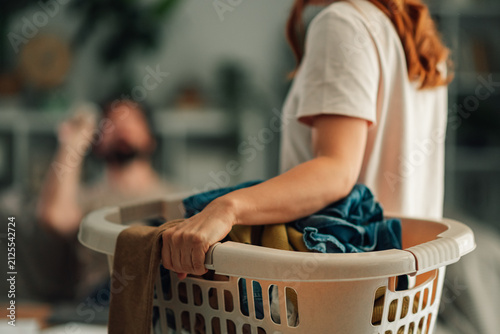 Woman carrying a full laundry basket with dirty clothes, performing household chores while partner rests on sofa