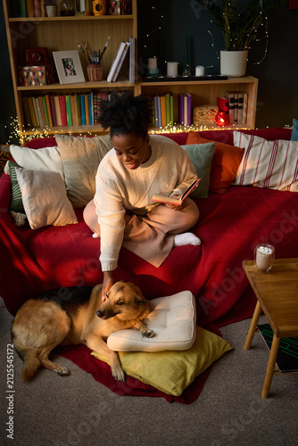 Photography Vertical image of young adult African American woman sitting on sofa, reading book and petting her dog