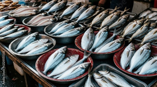 Rows of fresh fish rest in metal bowls at outdoor food market stall. Raw seafood harvest waits for sale on wooden counter table. Local vendor sells organic ocean catch to consumers. Traditional