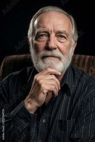 Portrait of serious older man with grey beard sitting in dark room. Senior male person looks at camera with thoughtful gaze. Mature guy rests chin on hand. Calm pensioner wears black striped shirt.