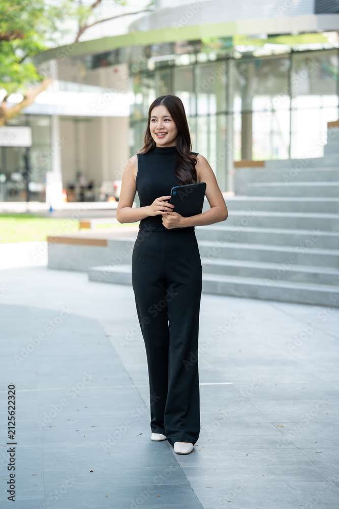 Fototapeta premium Young businesswoman standing outdoors with a tablet near a modern office building, smiling confidently in a bright urban setting.