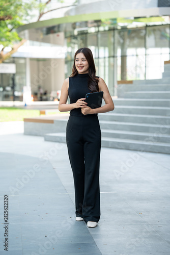 Young businesswoman standing outdoors with a tablet near a modern office building, smiling confidently in a bright urban setting.